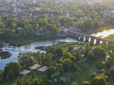 Skats no augšas uz Kuldīgas veco ķieģeļu tiltu, kas šķērso Ventas upi. Upes krastos izvietojusies pilsēta ar ēkām un baznīcas torni, ko ieskauj bagātīgi zaļi koki. Uz upes virsmas labajā pusē atspīd saule, un kreisajā krastā redzami smilšu laukumi un rotaļu laukums.