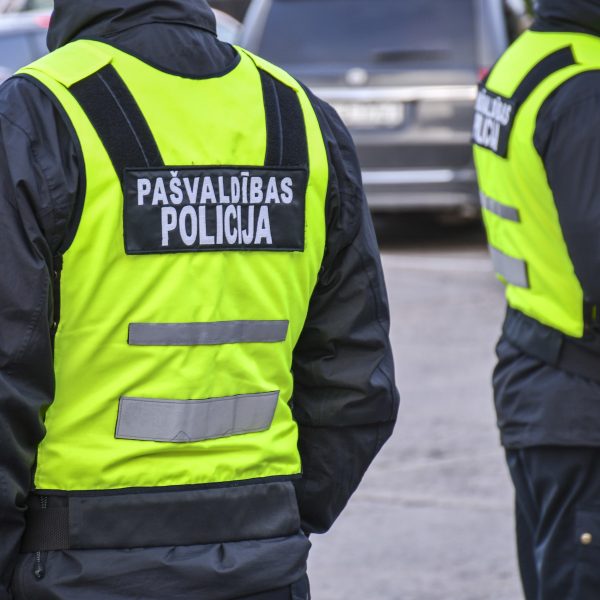 Divi pašvaldības policisti redzami no muguras, tērpušies melnās jakās un spilgti dzeltenās atstarojošās vestēs. Uz kreisā policista vestes ir uzraksts "PAŠVALDĪBAS POLICIJA".