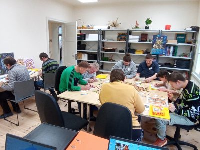 Vairāki cilvēki sēž pie diviem galdiem un ir iesaistīti radošā darbnīcā, zīmējot un krāsojot ar krāsu zīmuļiem. Fonā redzams plaukts ar grāmatām, mapēm un dažādiem dekoratīviem priekšmetiem, ieskaitot plakātu ar fantastiskas radības attēlu.