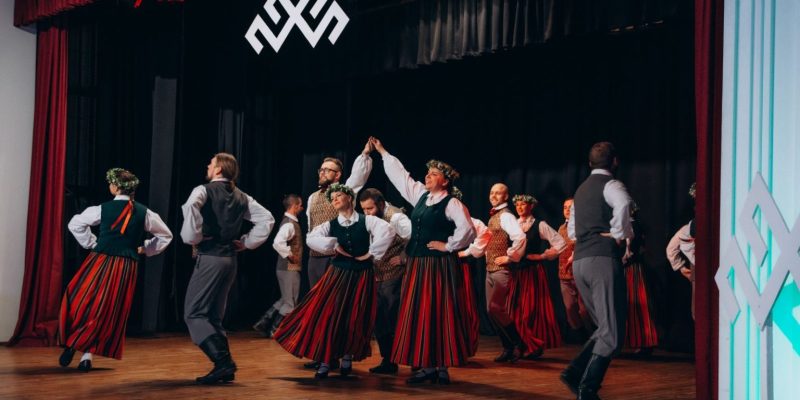 Folkloras dejotāji tradicionālos latviešu tērpos uzstājas uz skatuves. Sievietes valkā garus svītrainus svārkus, zaļas vestes un vainagus, vīrieši – pelēkas bikses un vestes. Aiz dejotājiem pie melnās sienas redzama liela balta Jumja zīme.