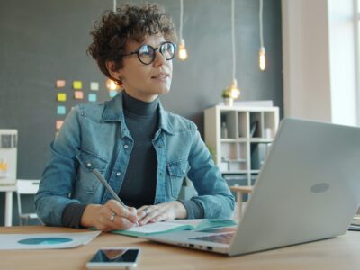 Pārdomās iegrimusi sieviete ar lokainiem matiem un brillēm sēž pie galda, rakstot piezīmju grāmatiņā ar pildspalvu, kamēr priekšā atvērts portatīvais dators modernā birojā.