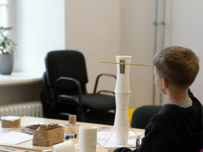 Zēns no muguras stāv pie galda un skatās uz no baltām papīra krūzītēm un koka nūjiņām izveidotu torni. Uz galda redzami citi rokdarbu materiāli.