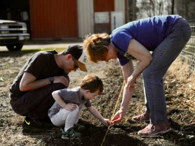 Trīs cilvēki – vīrietis, mazs bērns un sieviete – tupus stāda sēklas augsnē, izmantojot auklu kā vadlīniju. Sieviete rāda bērnam, kā to darīt. Saules apgaismotā fonā redzams sarkans šķūnis un kravas automašīna.